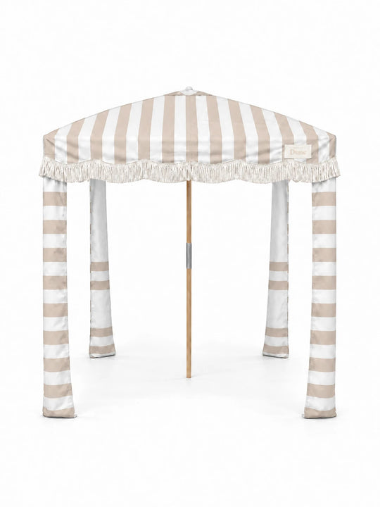 House of Dune Sand Drift beach cabana, front view on white: sand and white striped canopy, off-white fringe, striped fabric legs and a wooden centre pole.