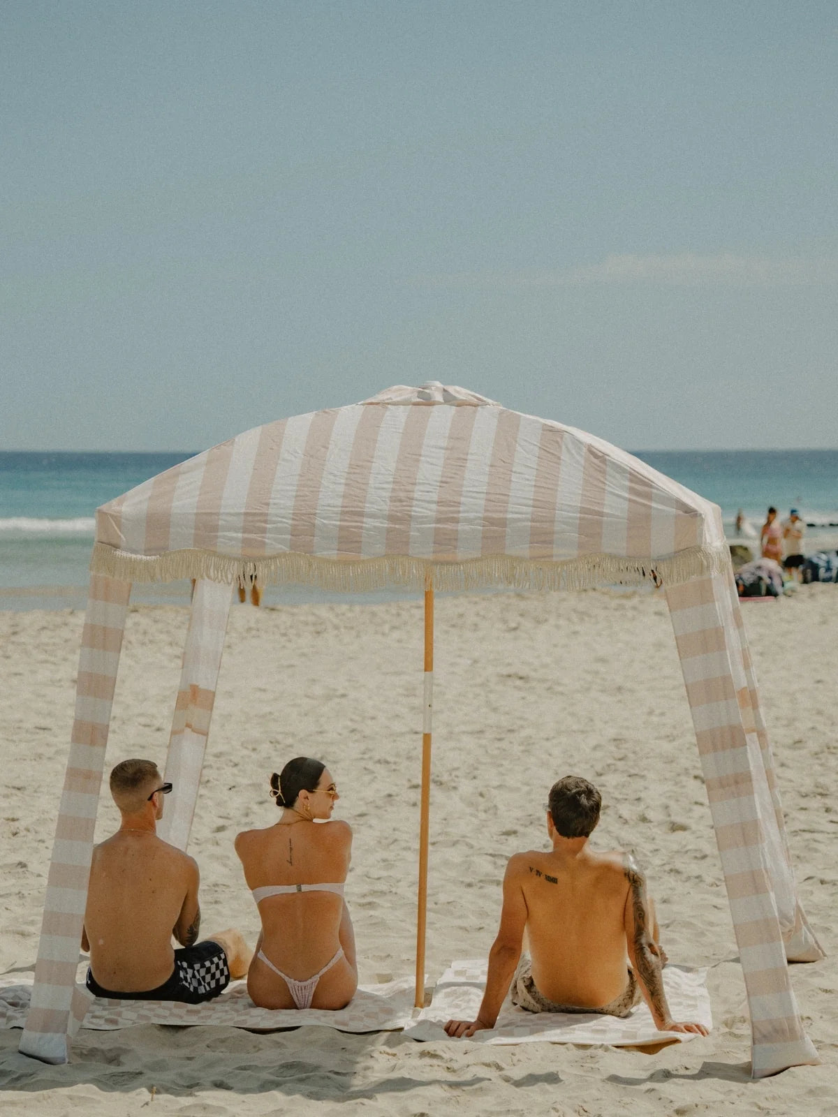 Three people relaxing under the House of Dune Sand Drift Cabana on a sandy beach, sitting on matching towels and enjoying a calm summer day by the ocean.