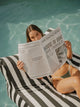 Woman lounging on the House of Dune Orca Stripe Floating Lounger while reading by the pool, highlighting the floating bean bag’s comfort and style.