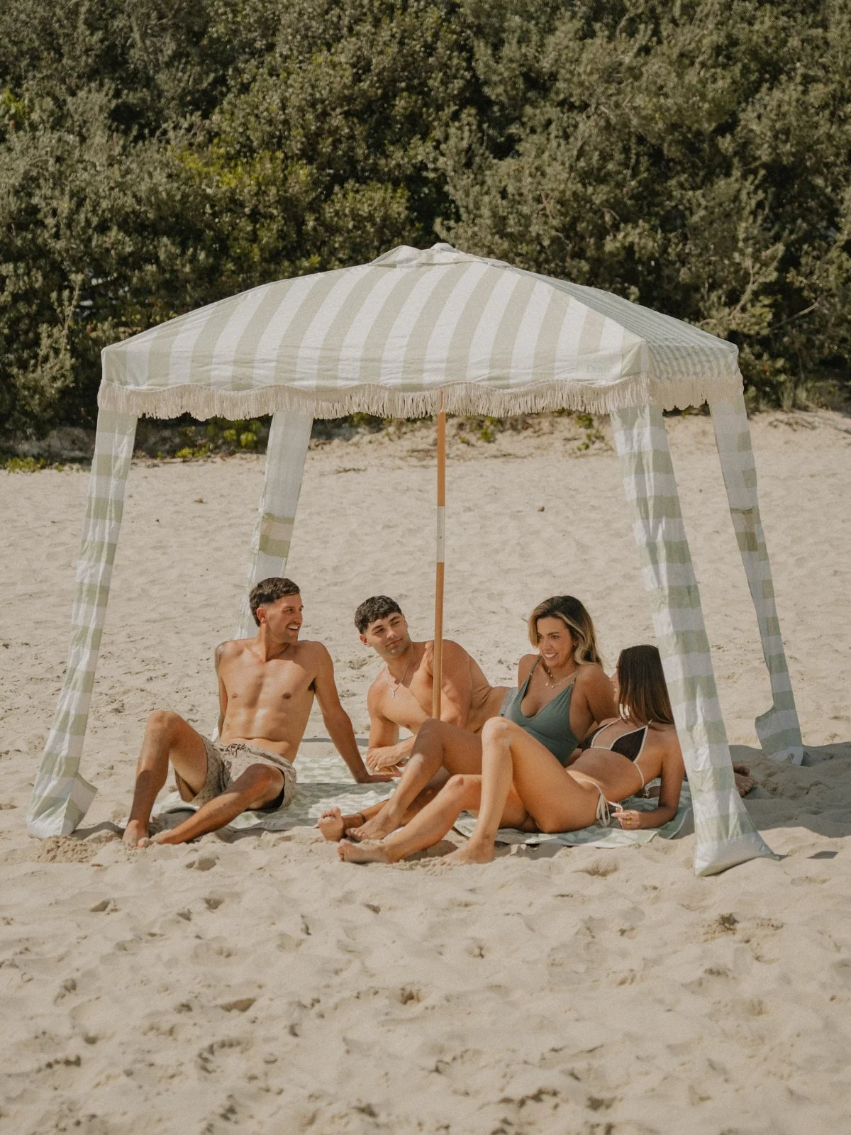 A group of friends gathered beneath the House of Dune Salted Sage Cabana enjoying a relaxed beach day, sitting on matching towels and chatting in the sun.