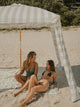 Two women unwind on checkered towels under the House of Dune Salted Sage Cabana, basking in the soft shade of its striped canopy for a serene seaside moment.