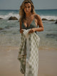 Woman in green swimsuit standing on the beach holding a Salted Sage checkered towel by House of Dune with waves behind her.