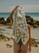 Back view of woman drying her hair with a Salted Sage checkered towel near ocean rocks, House of Dune coastal scene.
