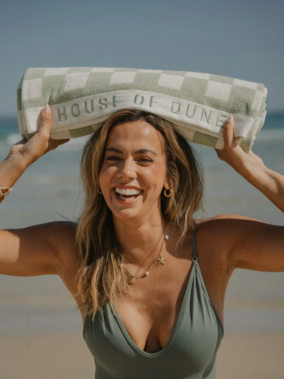 Smiling woman in green swimsuit holding a rolled Salted Sage checkered beach towel by House of Dune.
