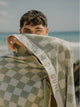 Man holding a Salted Sage checkered towel by House of Dune against a sunny coastal backdrop.