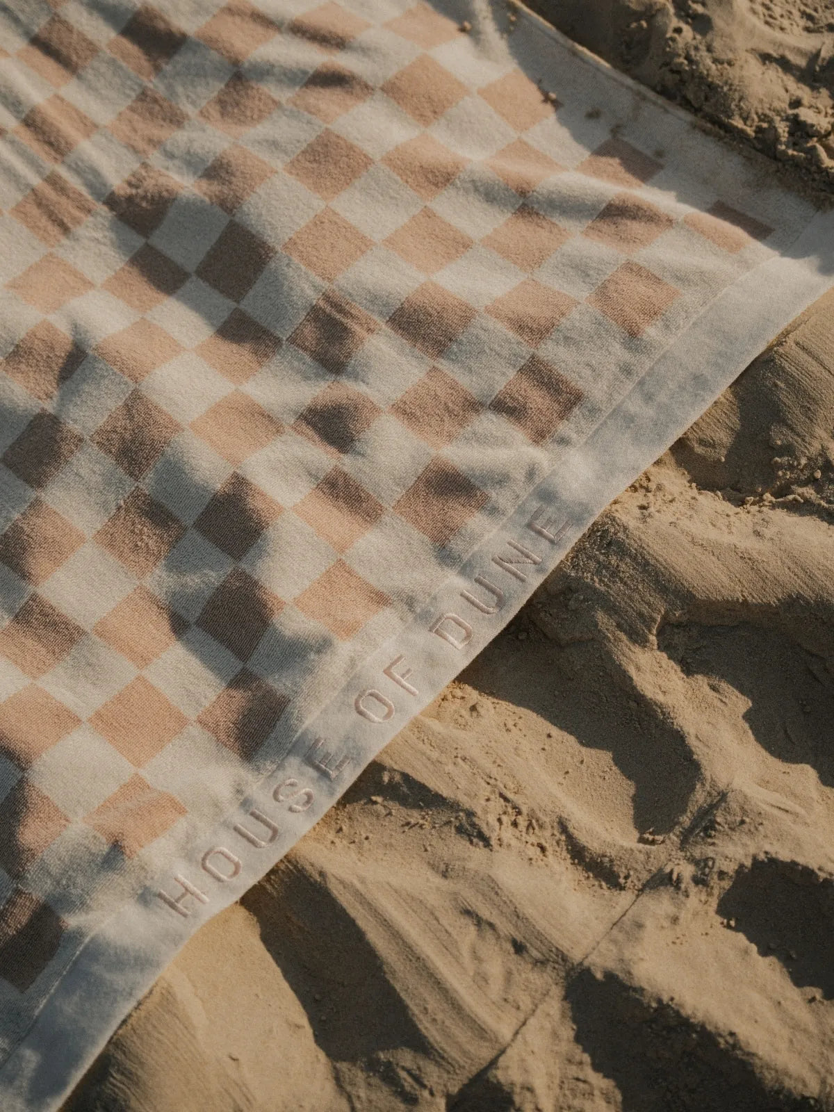 Close-up of Sand Drift checkered beach towel with embossed House of Dune logo resting on soft golden sand.