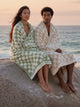 Two people wrapped in House of Dune checkered towels sitting on a rock at the beach during sunset, coastal tones.