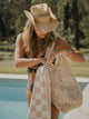 Woman in swimsuit and straw hat packing a Sand Drift checkered towel into a woven beach bag by House of Dune.