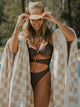 Smiling woman in brown bikini and hat wrapped in a Sand Drift checkered towel by House of Dune beside the pool.
