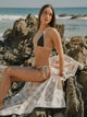 Woman in brown bikini sitting on coastal rocks with Sand Drift checkered towel draped beside her, House of Dune.