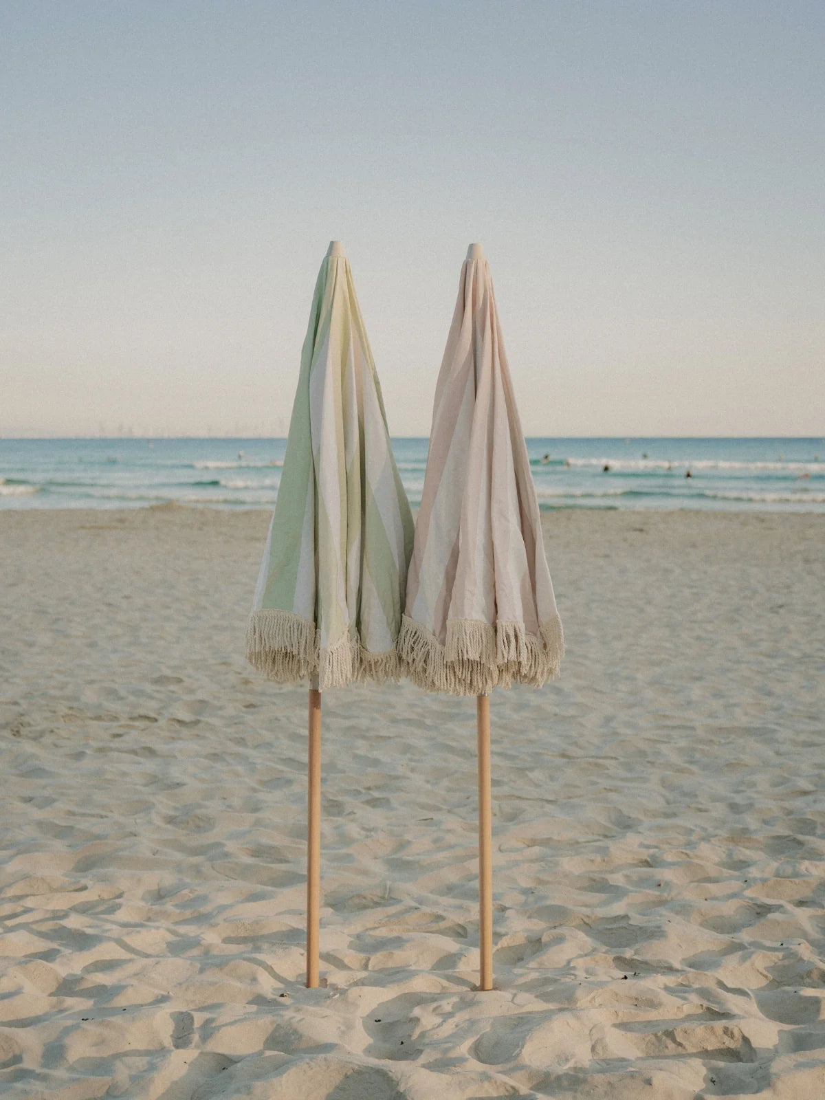 Salted Sage and Sand Drift beach umbrellas from House of Dune folded side by side on the sand near the ocean.
