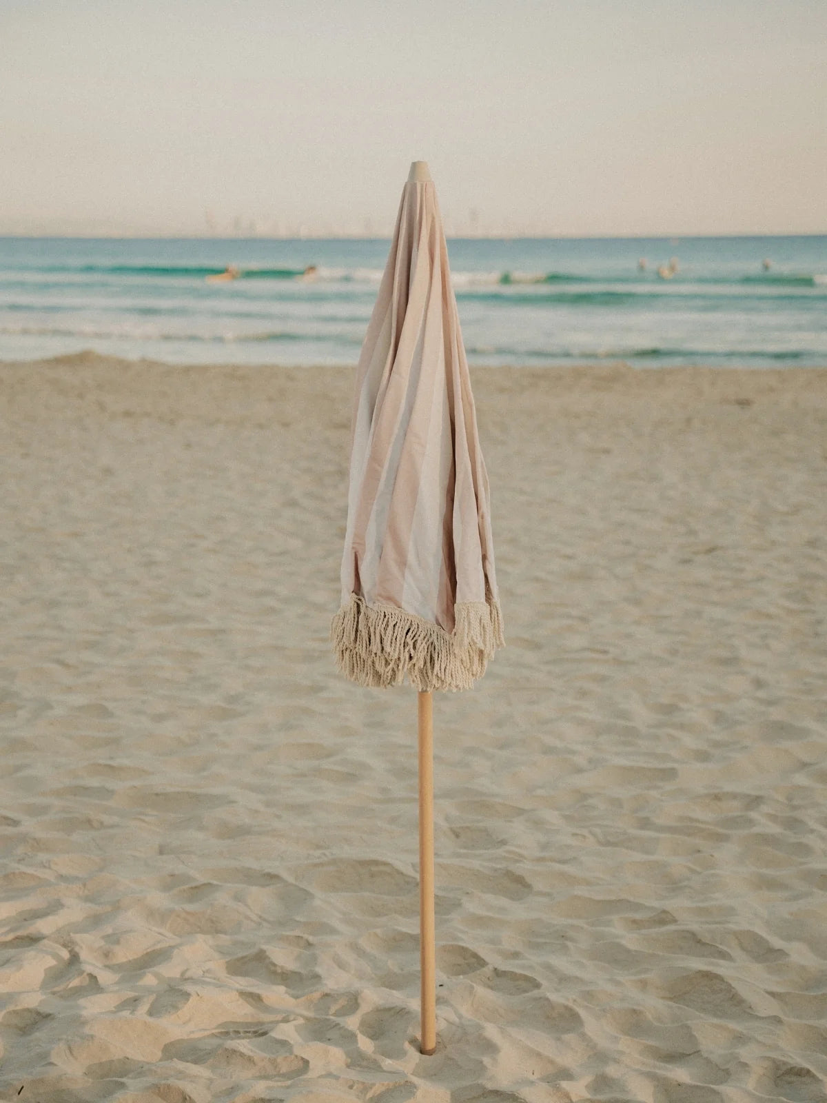 Sand Drift beach umbrella folded upright in the sand with ocean waves in the background, by House of Dune.