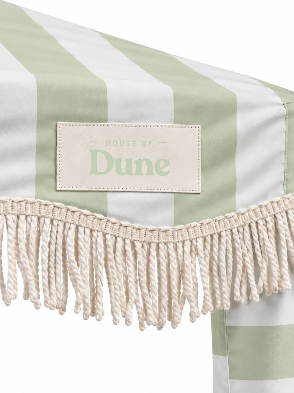 Close-up of the House of Dune Salted Sage cabana corner: stitched House of Dune badge on the sage striped canopy above the twisted off-white cotton fringe.