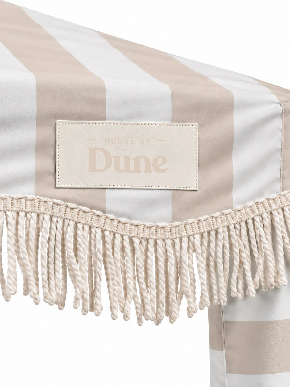 Close-up of the House of Dune Sand Drift cabana corner: stitched House of Dune badge on the sand striped canopy above the twisted off-white cotton fringe.