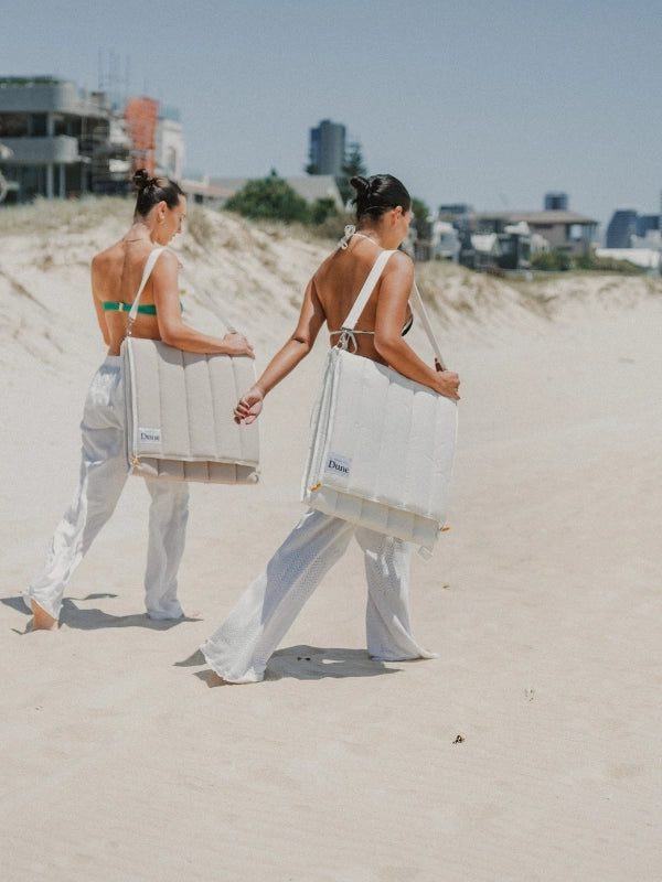 Two women carrying folded House of Dune beach loungers over their shoulders by the carry strap, walking over the dunes.