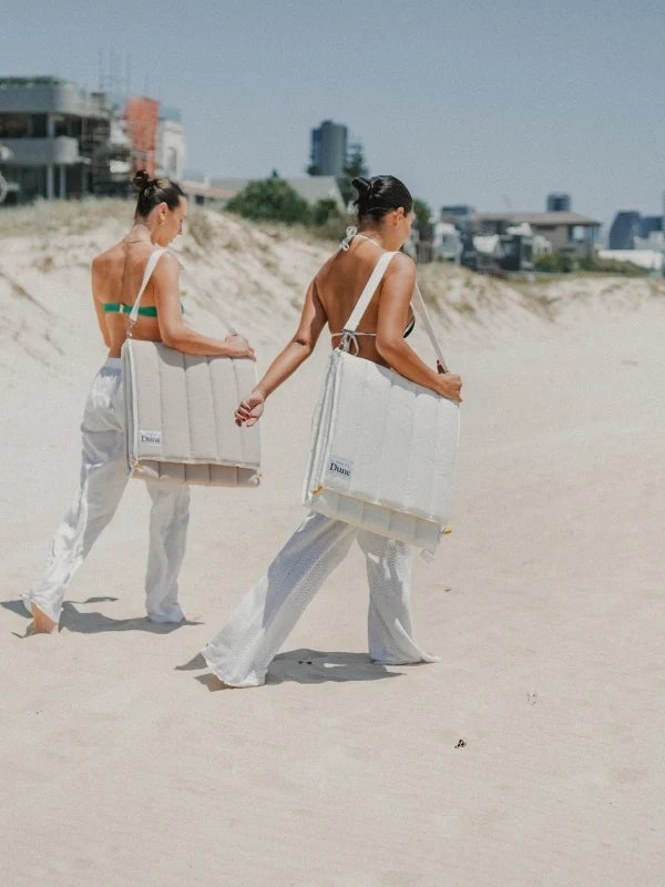 Two women carrying folded House of Dune beach loungers over their shoulders by the carry strap, walking over the dunes.