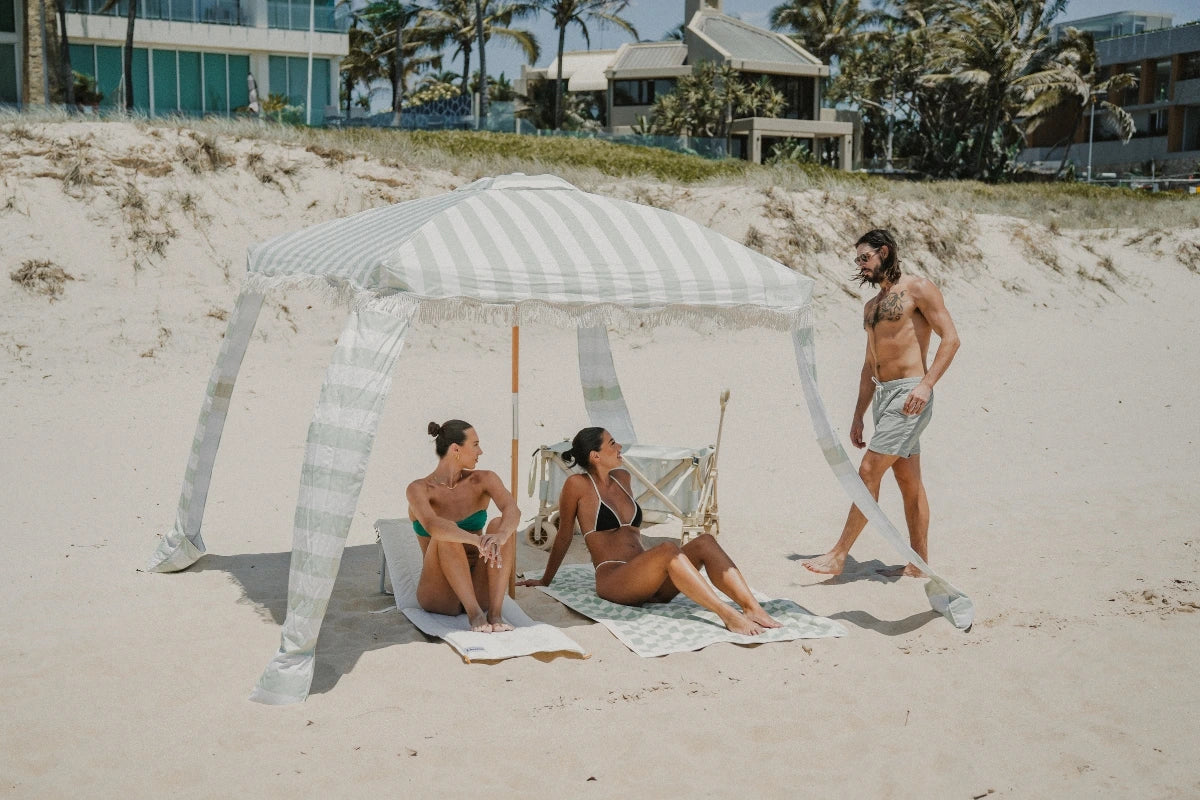 House of Dune Oyster Tide and Sand Drift beach loungers made from recycled RPET, styled beneath a striped beach umbrella for shaded coastal lounging on soft sand.