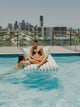 Two women relaxing on a House of Dune Salted Sage floating lounger in a rooftop pool with city views.