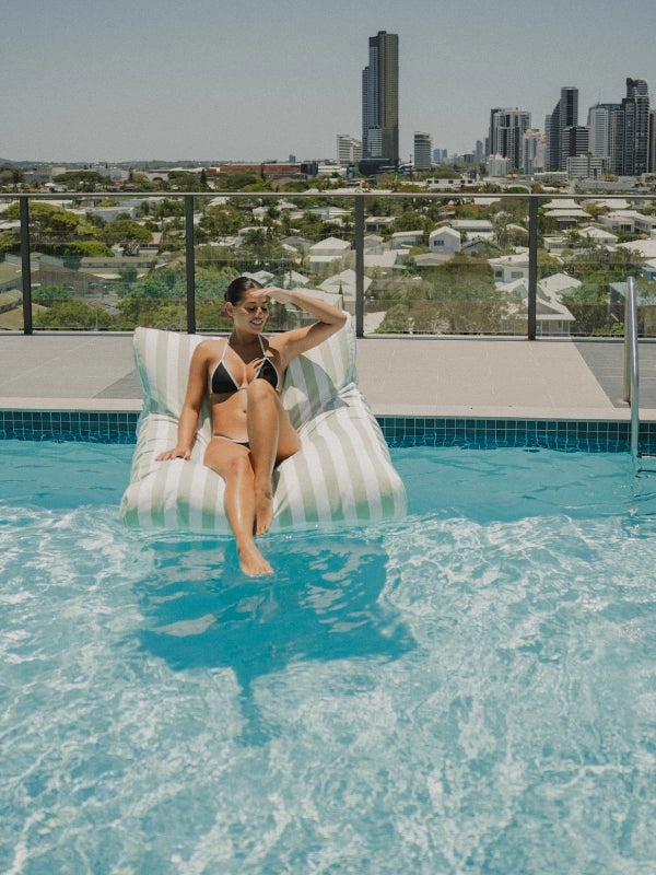 Woman lounging on the House of Dune Salted Sage floating lounger in a rooftop pool, city skyline behind.