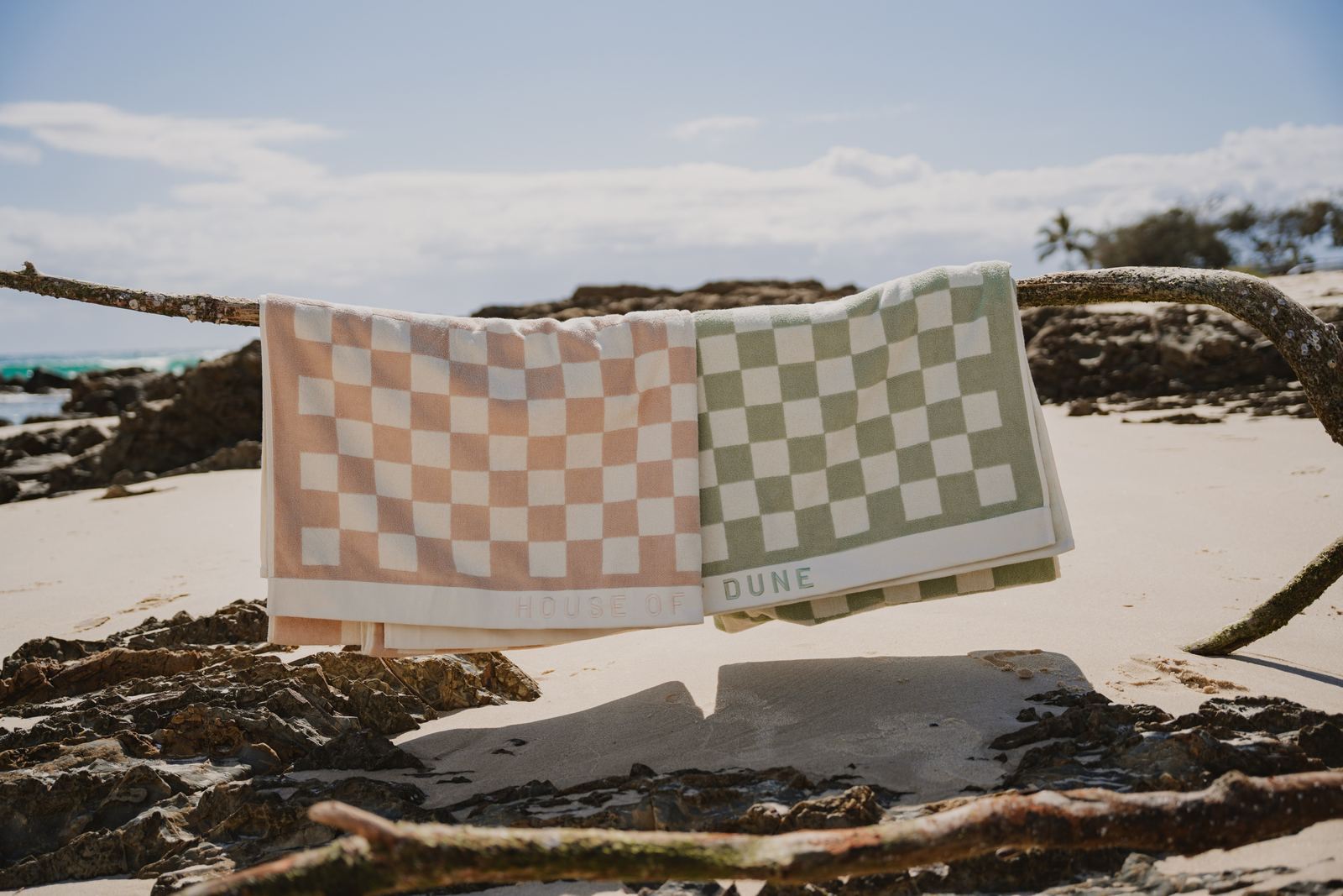 Two House of Dune beach towels, Sand Drift and Salted Sage, drying on driftwood on the sand