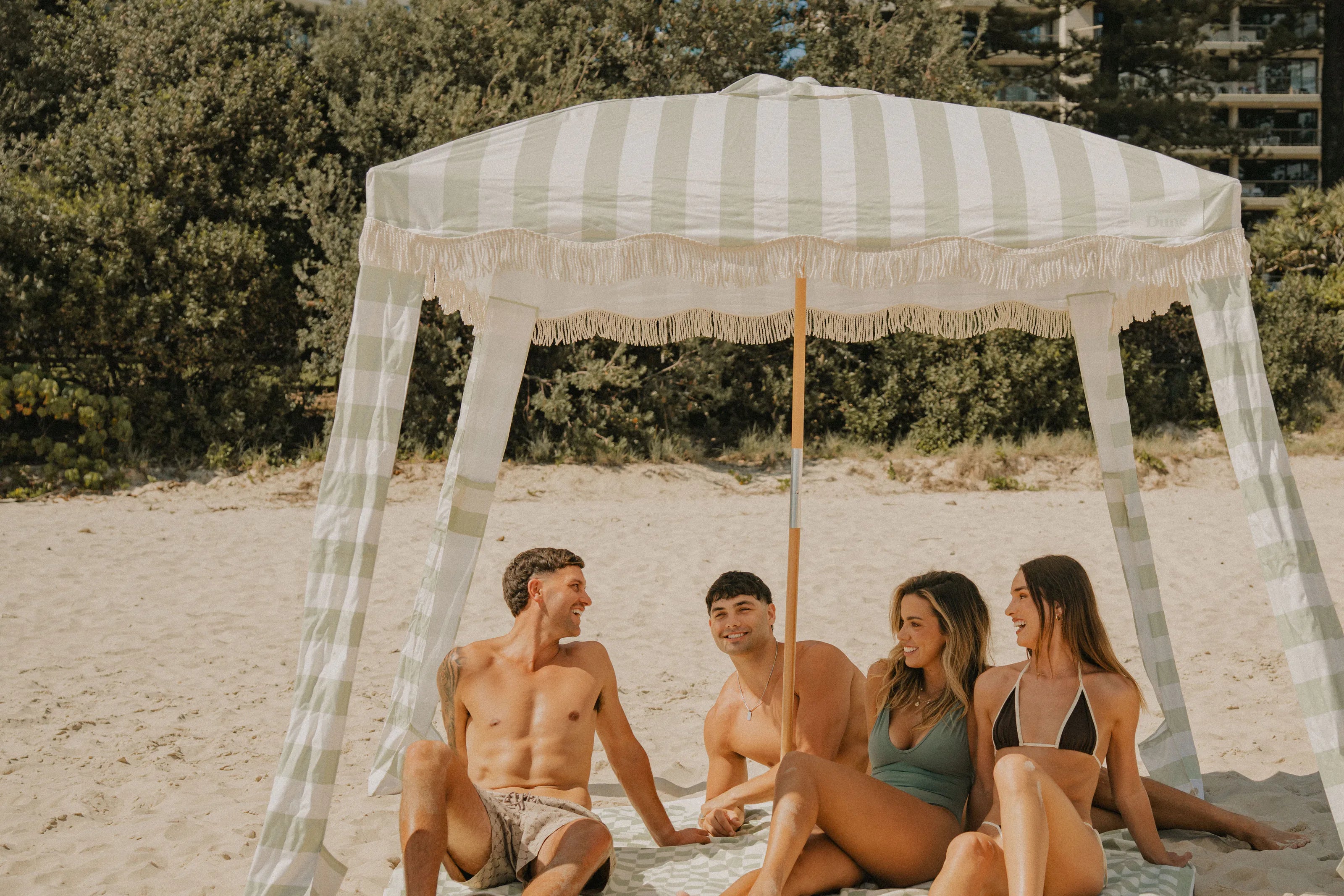 A Salted Sage striped beach cabana on the sand, friends resting in its shade