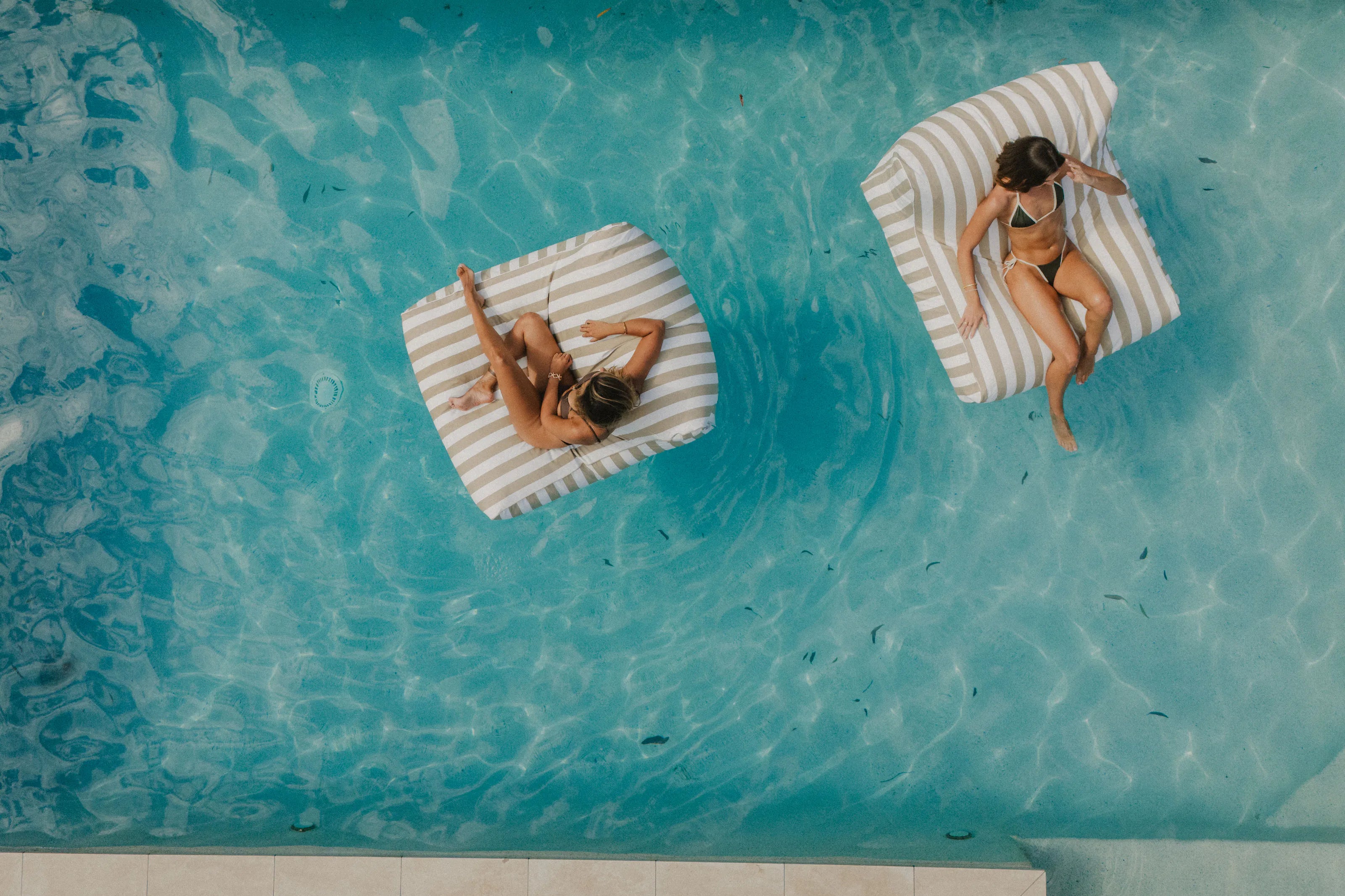 Aerial view of two Sand Drift floating loungers, one person on each, in a turquoise pool