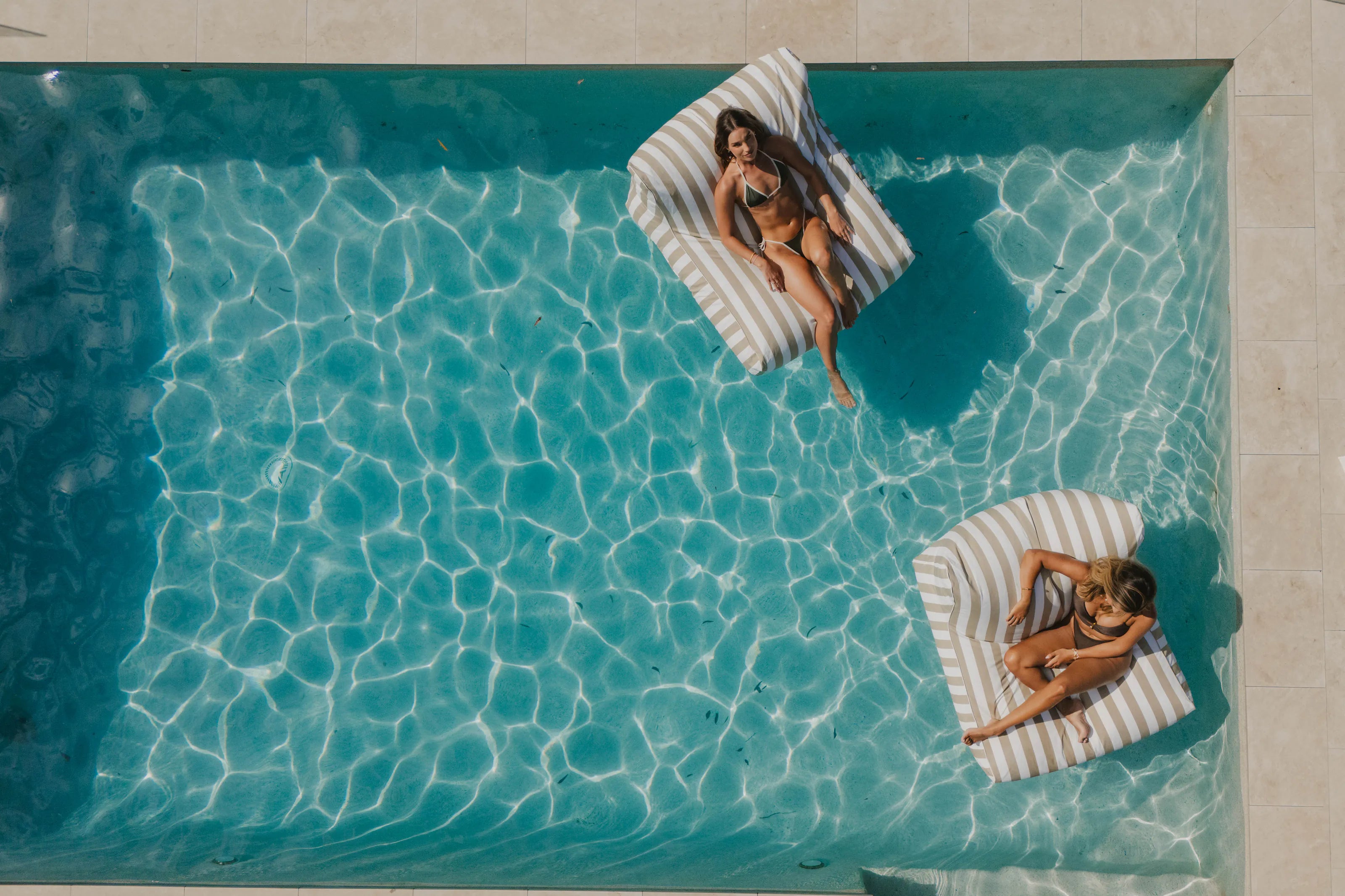 Two people relaxing on striped floating loungers in a turquoise pool, seen from above